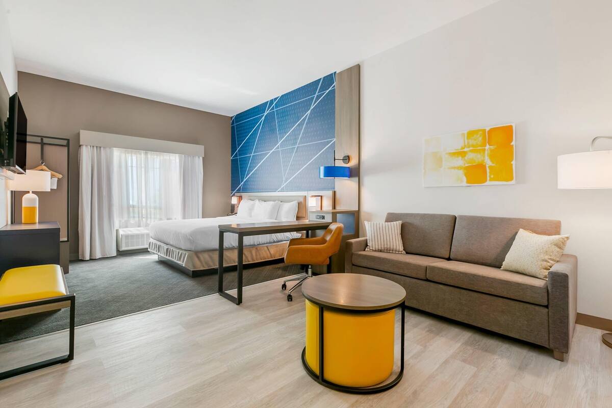 A modern hotel room with a bed, a sofa, a desk, and a chair. There is a large window with curtains, a wall-mounted TV, and artwork on the walls.