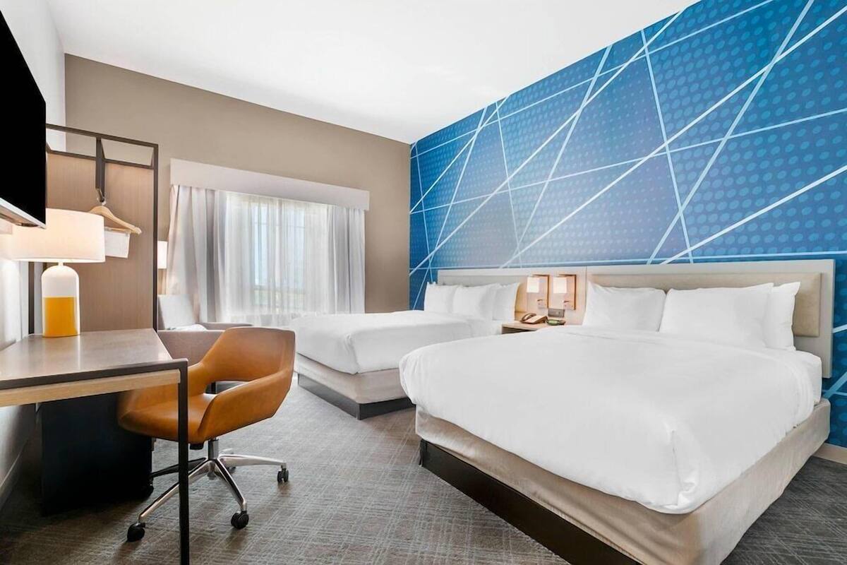 A hotel room with two beds, a desk, a chair, and a large window with curtains.