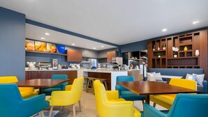 Un restaurant moderne avec des chaises jaunes et turquoise, un îlot central doté d’une machine à café et un mur en bois avec des étagères.