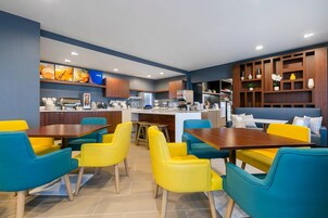 A modern restaurant with yellow and teal chairs, a central island with a coffee machine, and a wooden wall with shelves.