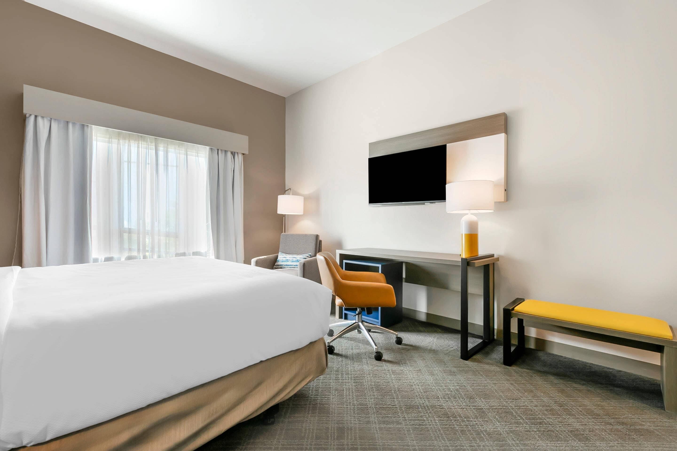 A hotel room with a large bed, a desk with a chair, a TV mounted on the wall, and a bench.