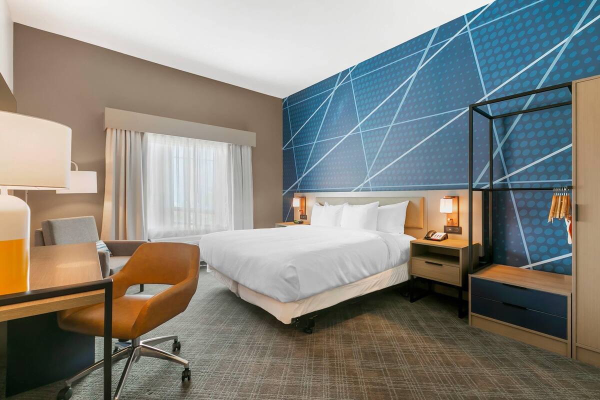 A hotel room with a large bed, a desk with a chair, a nightstand with a lamp, and a wardrobe.