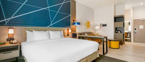 A modern hotel room with a large bed, bedside tables, a desk, and a kitchenette.