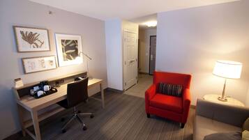 A hotel room with a desk, a red armchair, a grey sofa, a lamp, and framed botanical prints on the wall.