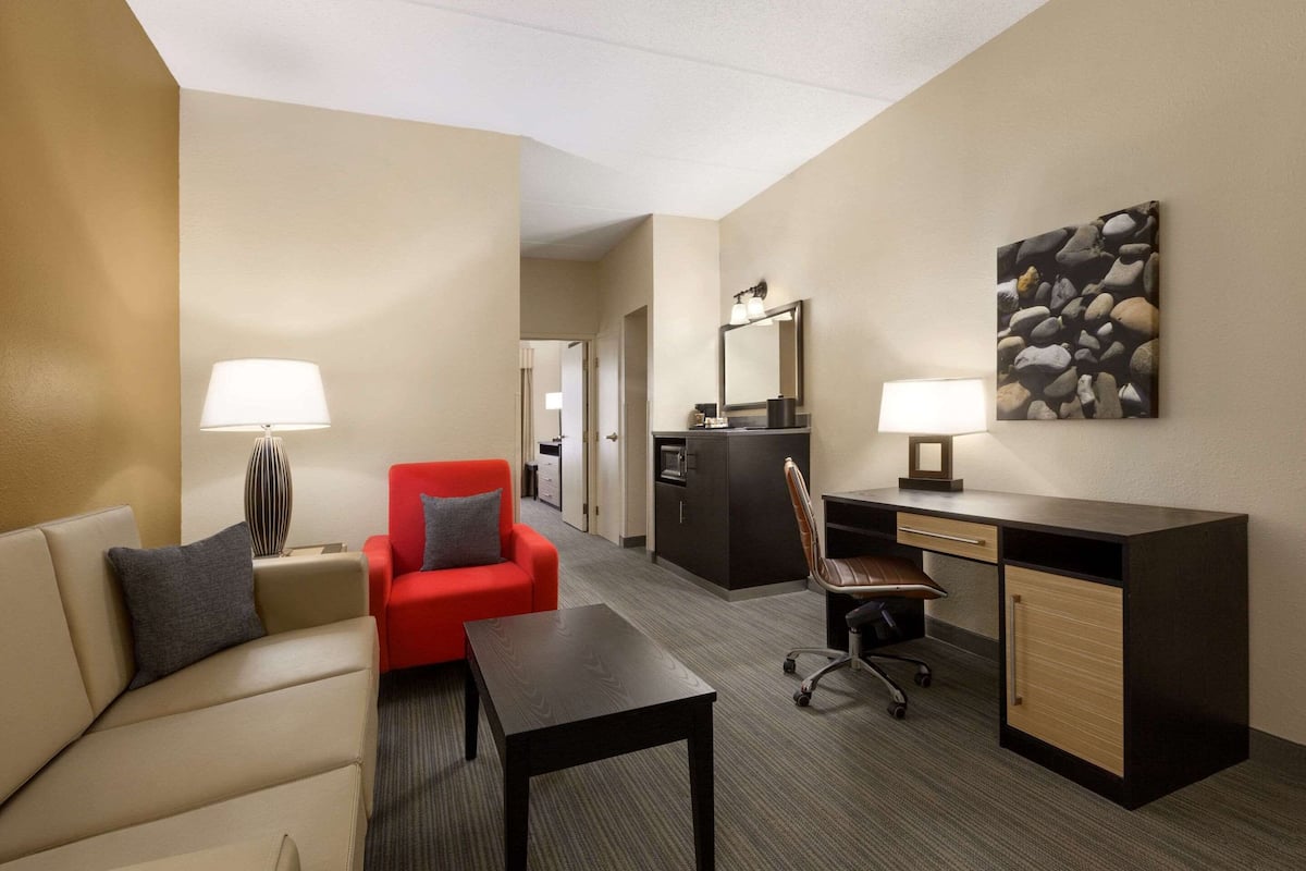 Una habitación de hotel con un sofá, un sillón rojo, un escritorio, una silla, una lámpara, un espejo y un cuadro en la pared.