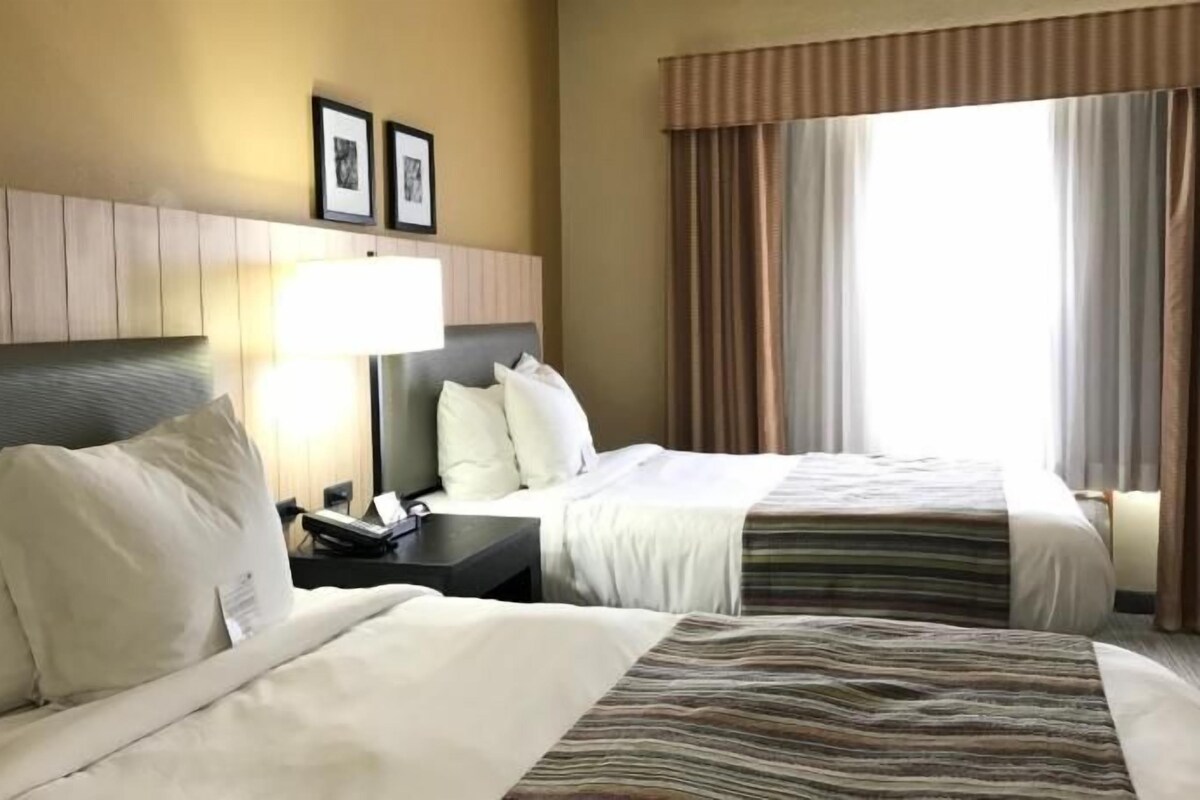 A hotel room with two beds, a nightstand, a lamp, and a window with curtains.