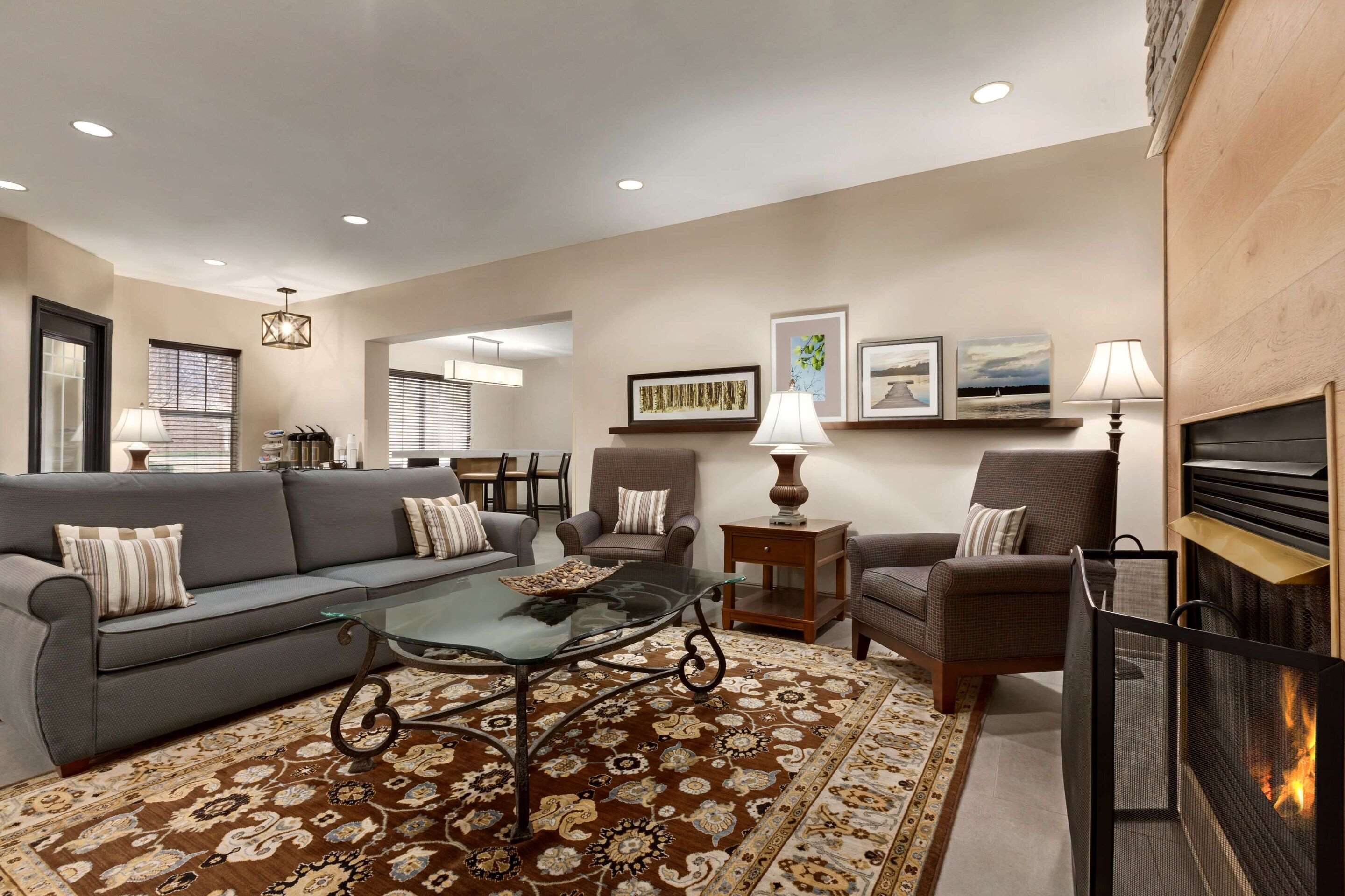 A living room with a sofa, chairs, a glass coffee table, a patterned rug, a fireplace, and wall-mounted artwork.