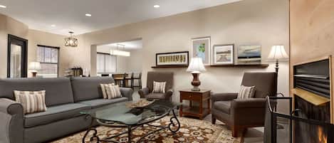 A living room with a sofa, chairs, a glass coffee table, a patterned rug, a fireplace, and wall-mounted artwork.