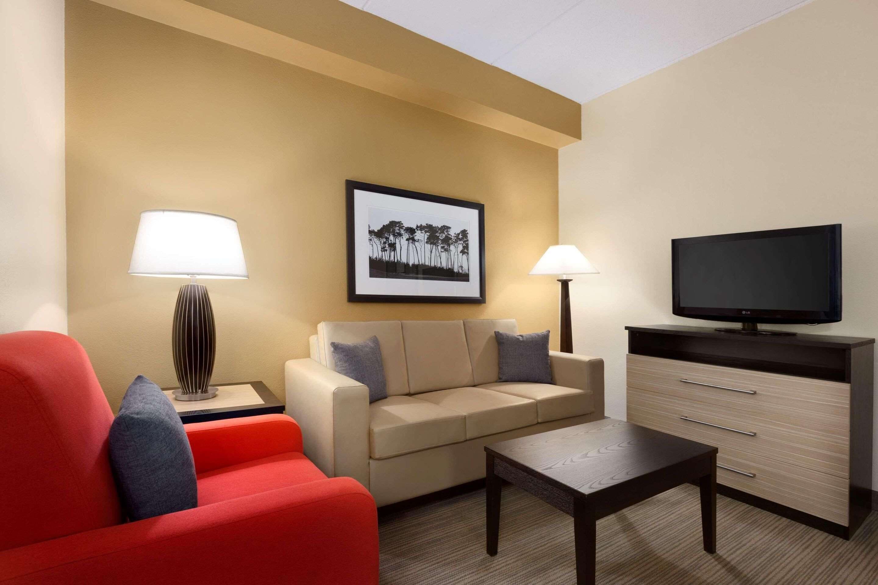 Quarto de hotel com um sofá bege, uma poltrona vermelha, uma mesa de centro, uma TV sobre um suporte e duas luminárias de chão.