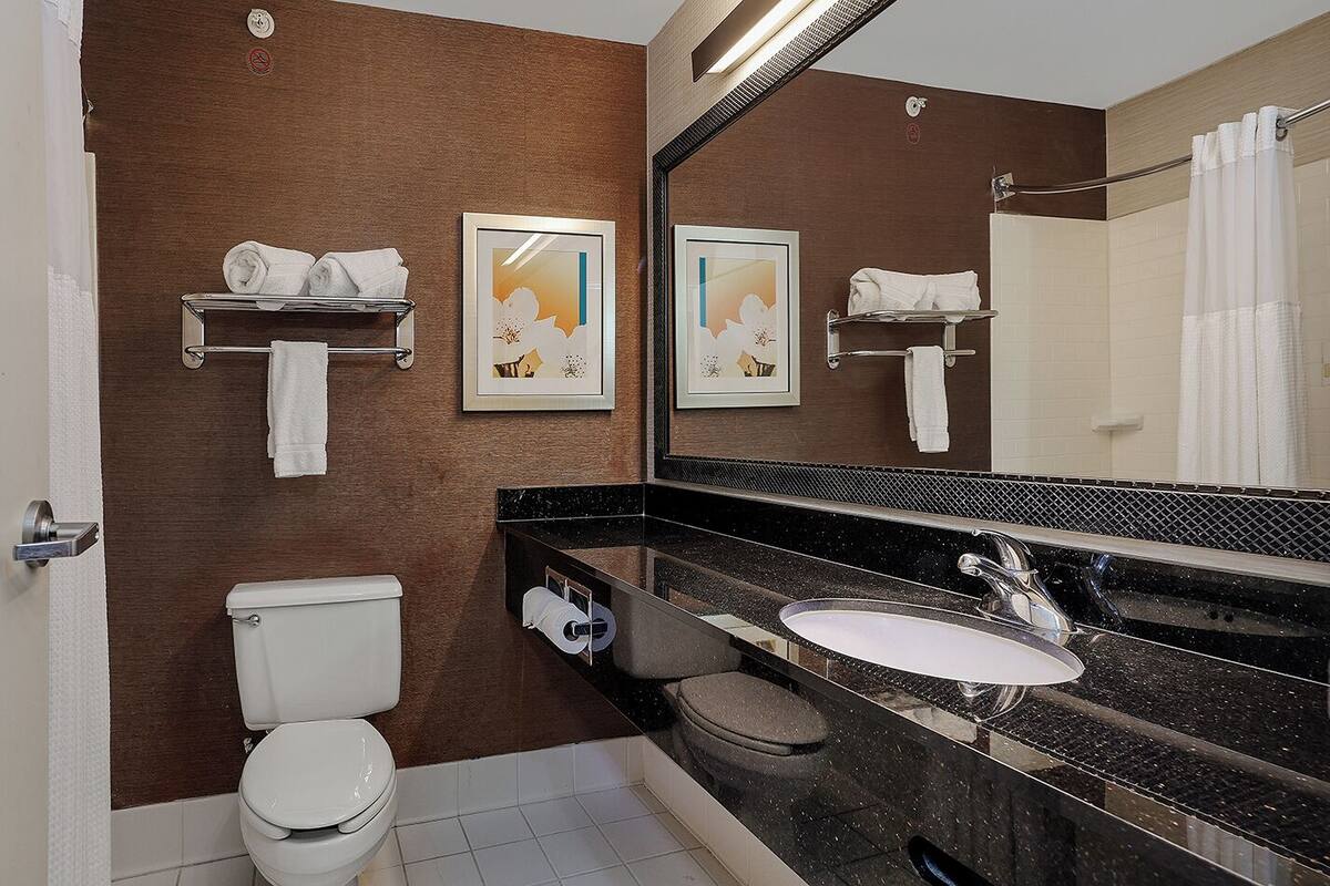 A modern bathroom with a toilet, sink, and large mirror. There are towels on the wall and a picture frame.