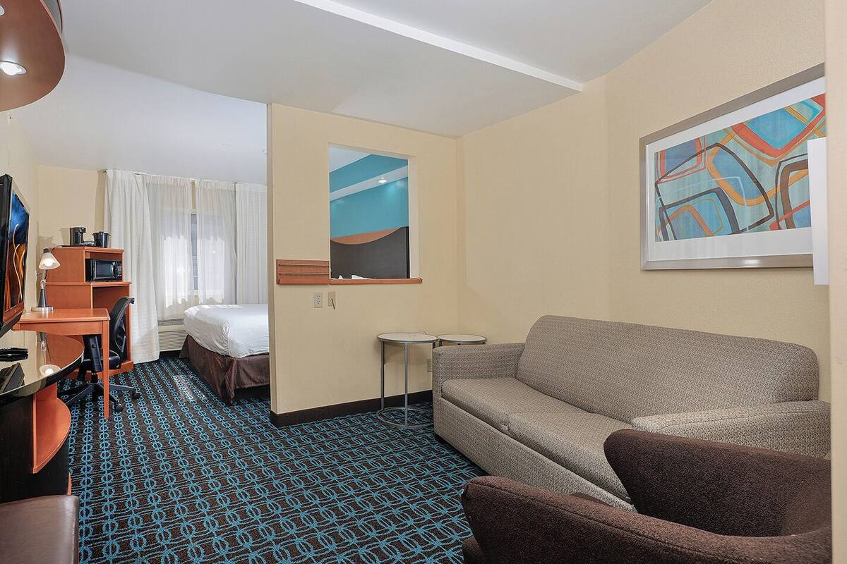 A hotel room with a bed, a sofa, a desk with a lamp, a small table, and a painting on the wall.