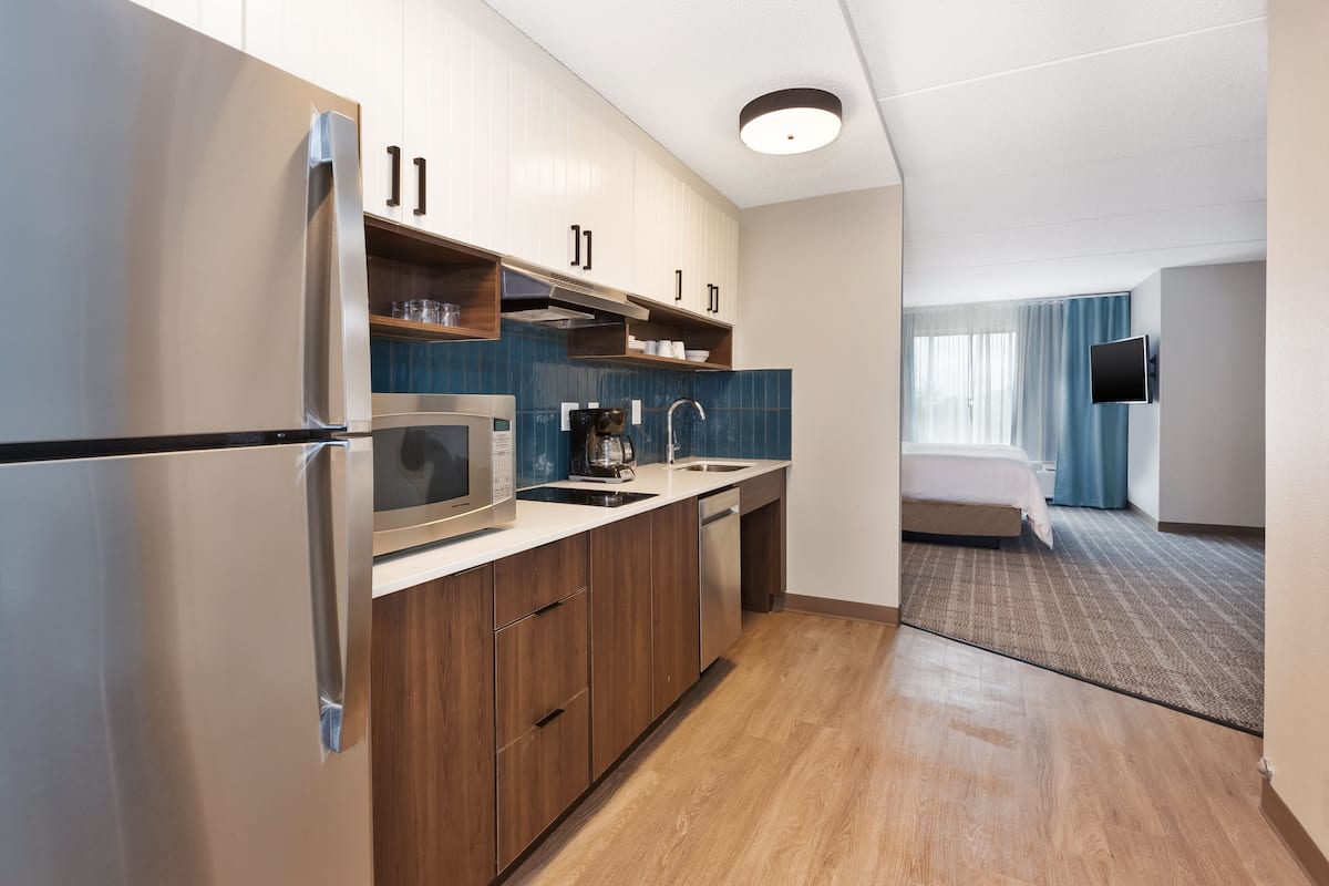 A modern kitchen with stainless steel appliances, wooden cabinets, and a view into a bedroom with a bed and curtains.