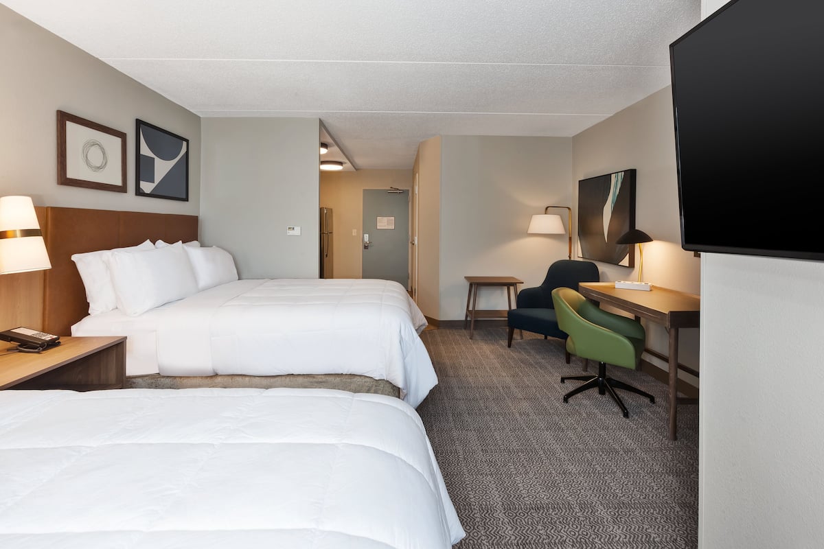 A hotel room with two beds, a desk, a chair, a TV, and a lamp.
