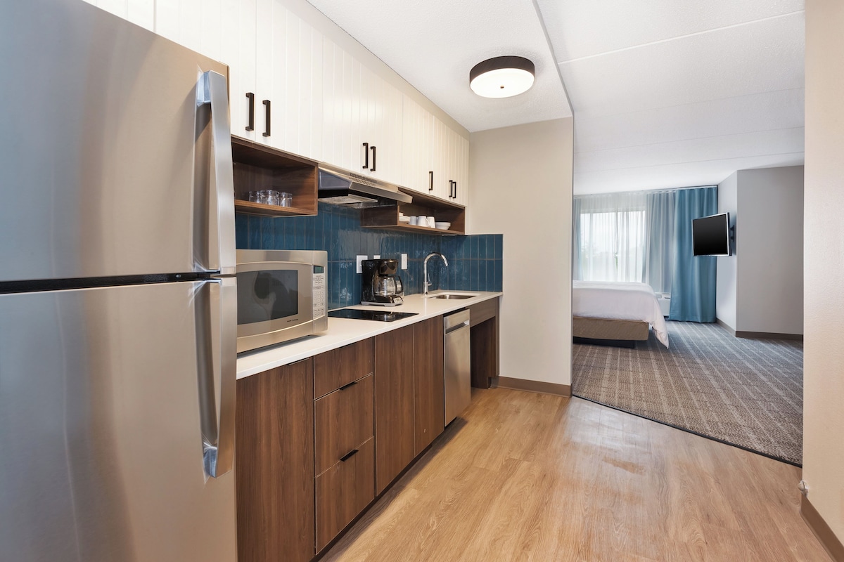 Una cocina moderna con electrodomésticos de acero inoxidable, armarios de madera y vistas a un dormitorio con cama y cortinas.