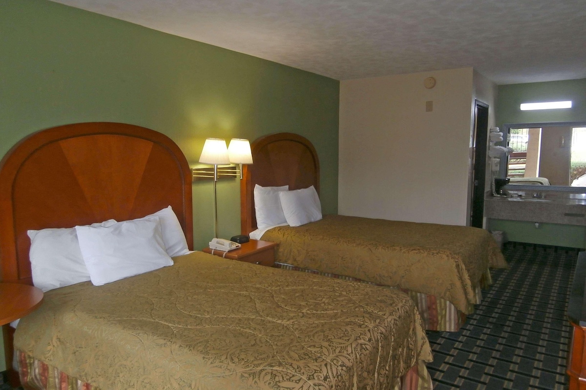 Two single beds with wooden headboards, a nightstand with a lamp, a small table, and a bathroom with a mirror and sink.