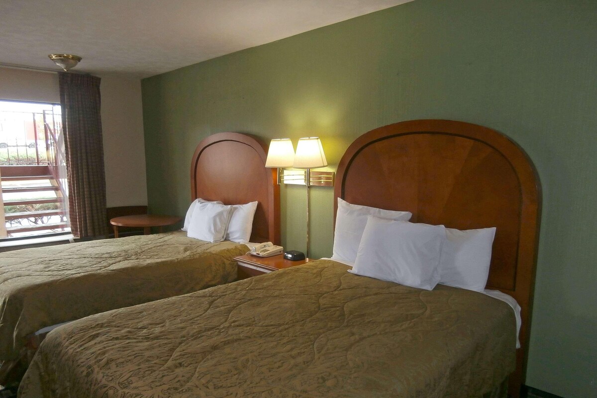 A hotel room with two beds, a nightstand with a lamp, a telephone, and a view of the outside.