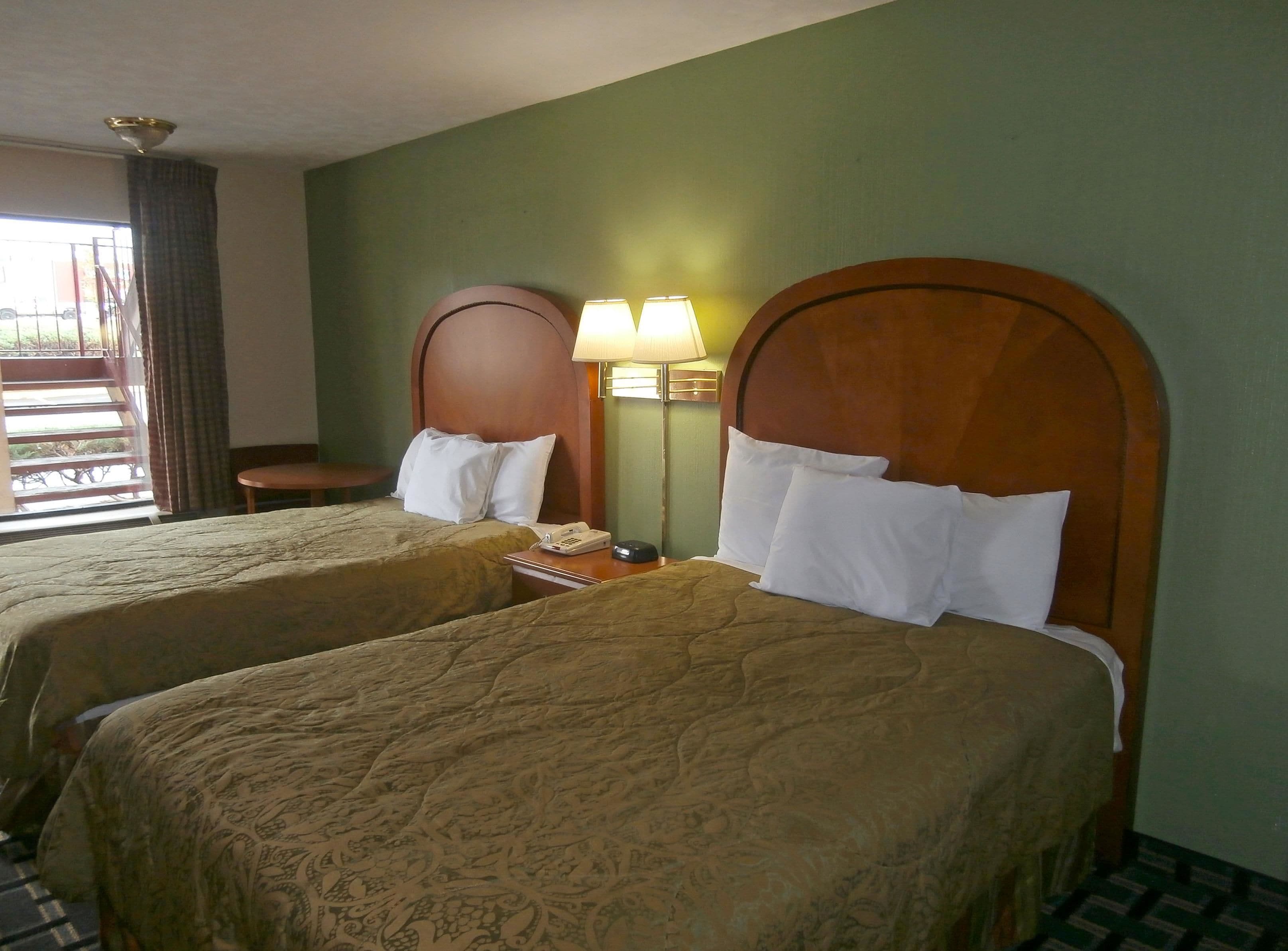 A hotel room with two beds, each with a wooden headboard and white pillows. There is a nightstand with a lamp and a telephone on one side.