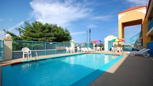 A swimming pool with lounge chairs and umbrellas, surrounded by a fence and a building with a red roof.