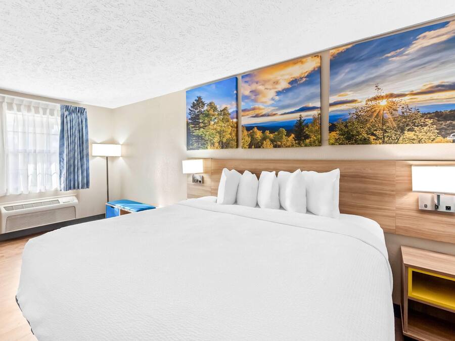 Habitación de hotel con una cama grande, dos lámparas de pared, un ventanal con cortinas y un cuadro de un paisaje al atardecer.