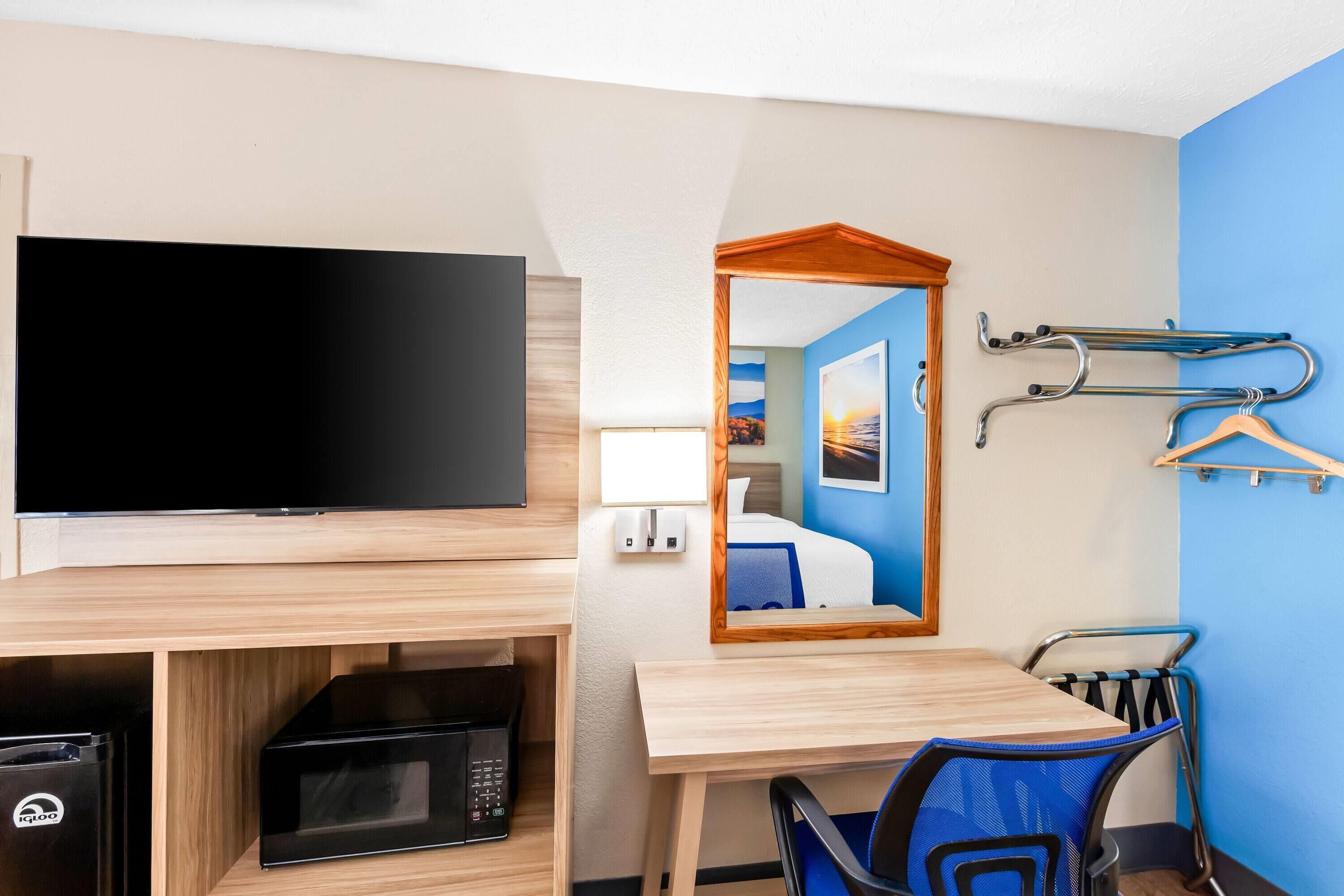 A hotel room with a flat-screen TV, a microwave, a wooden desk, a chair, a mirror, and a coat rack with hangers.