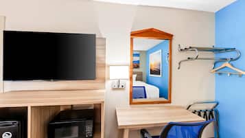A hotel room with a flat-screen TV, a microwave, a wooden desk, a chair, a mirror, and a coat rack with hangers.