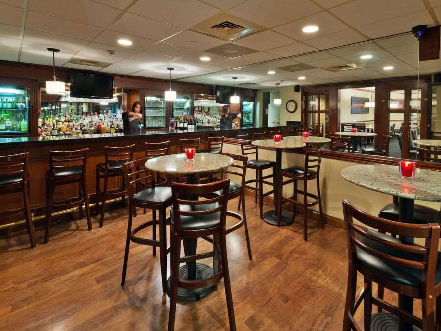 A bar area with multiple round tables and chairs, a bar counter with a variety of bottles, and a television mounted on the wall.