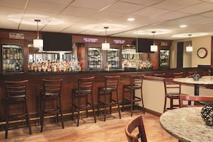 Zona de bar con varios taburetes, una barra con botellas y un reloj en la pared.