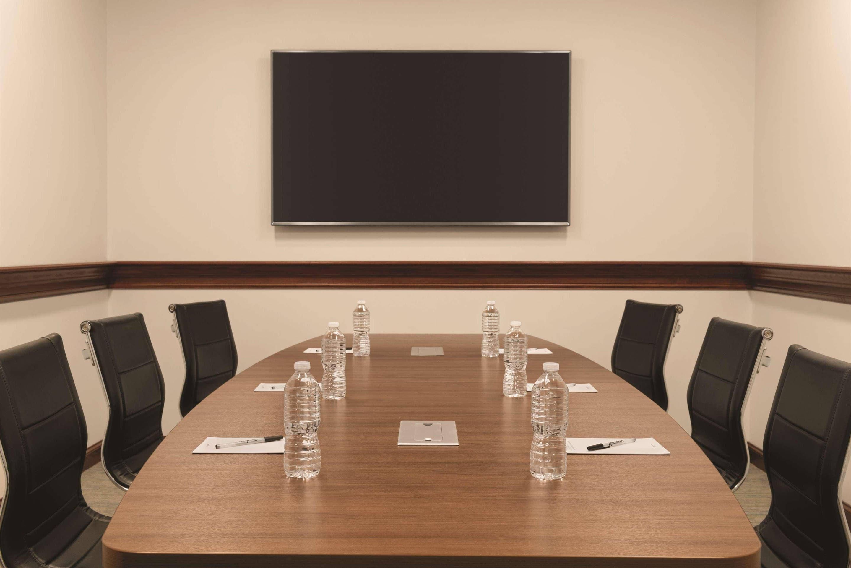 Un salón de conferencias con una larga mesa, sillas negras, botellas de agua y una pantalla de proyector.