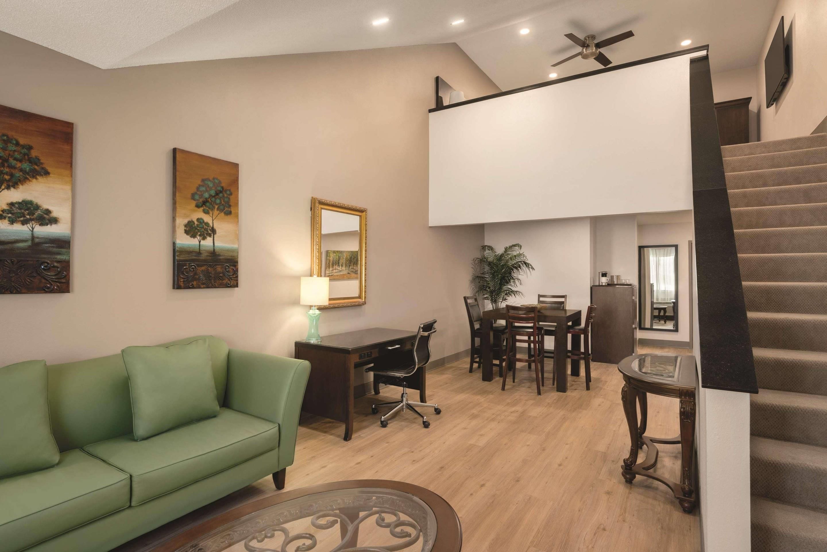A modern living room with a green sofa, a dining area with a table and chairs, a desk with a lamp, and a staircase.