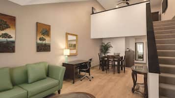 A modern living room with a green sofa, a dining area with a table and chairs, a desk with a lamp, and a staircase.