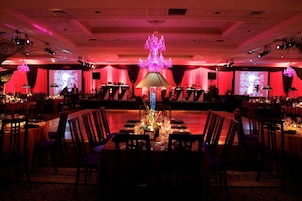 Un salón de banquetes con mesas dispuestas para un evento formal, un escenario con equipo musical y una araña.