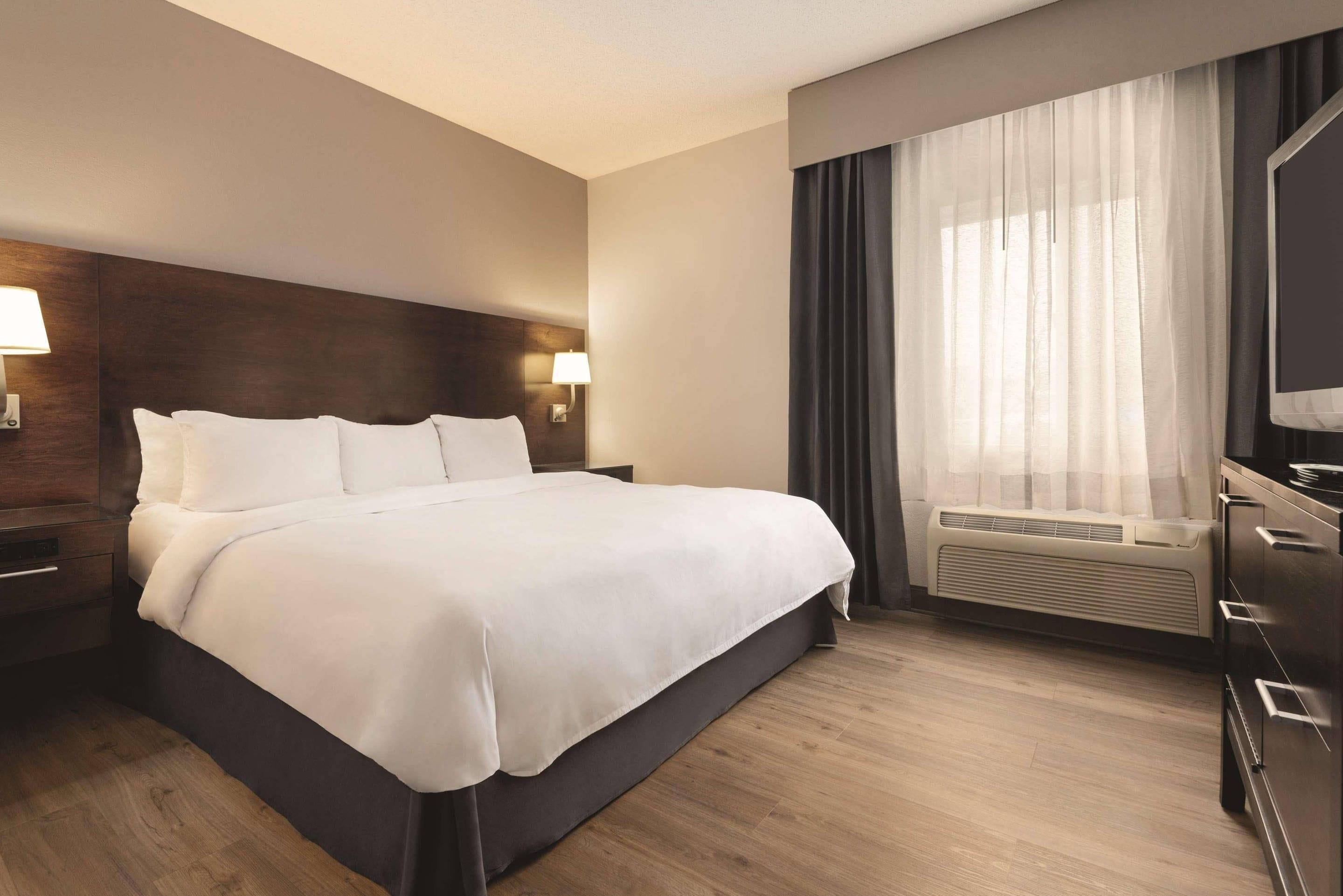 Habitación de hotel con una cama grande, cabecera de madera, mesita de noche con lámpara, cómoda y ventana con cortinas.