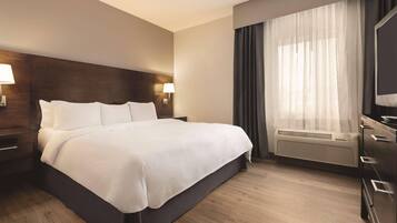 Habitación de hotel con una cama grande, cabecera de madera, mesita de noche con lámpara, cómoda y ventana con cortinas.