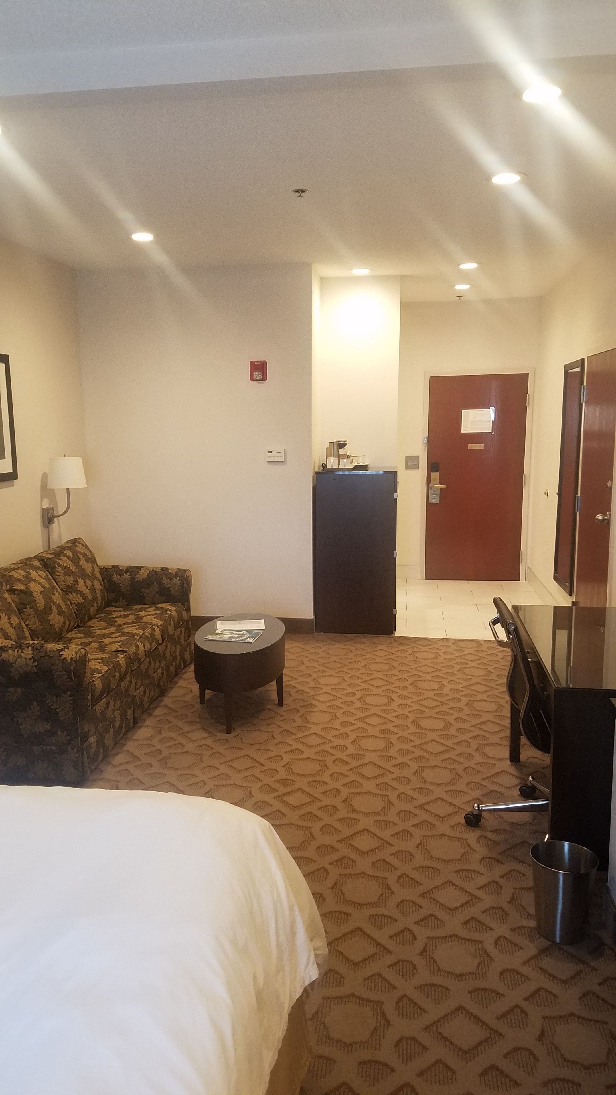A hotel room with a bed, a sofa, a small table, a desk, a mirror, and a door.