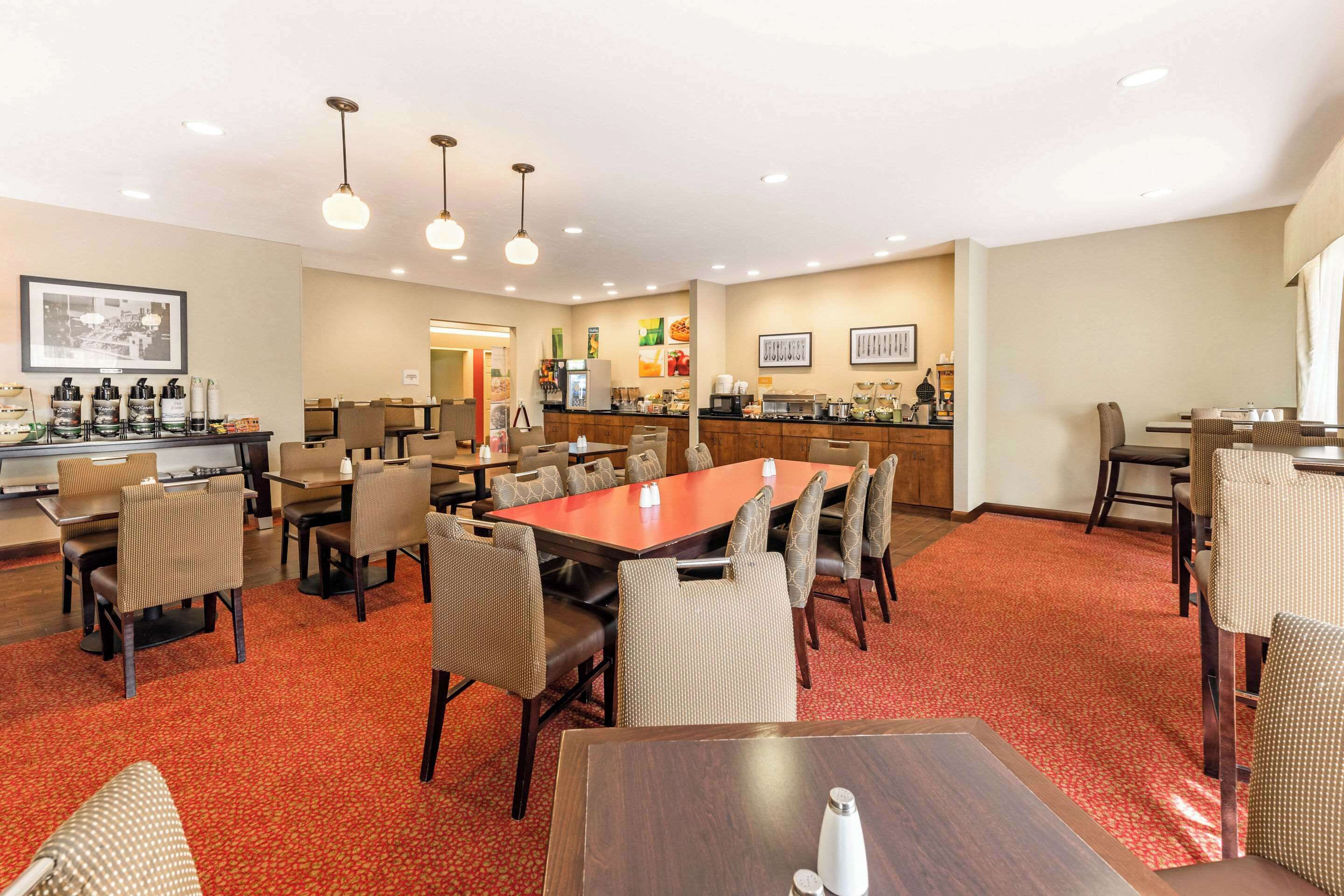 A hotel dining area with multiple tables and chairs, a kitchen in the background, and a red carpeted floor.