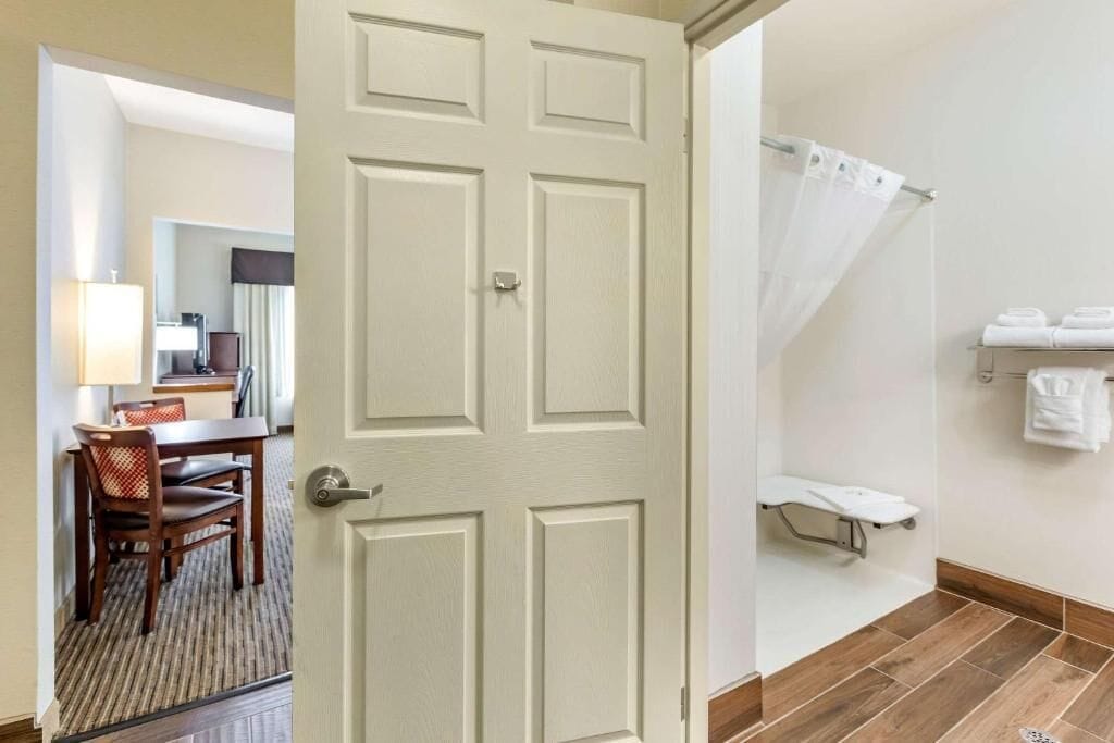 A hotel room with a closed door, a bathroom with a shower, and a dining area with a table and chairs.