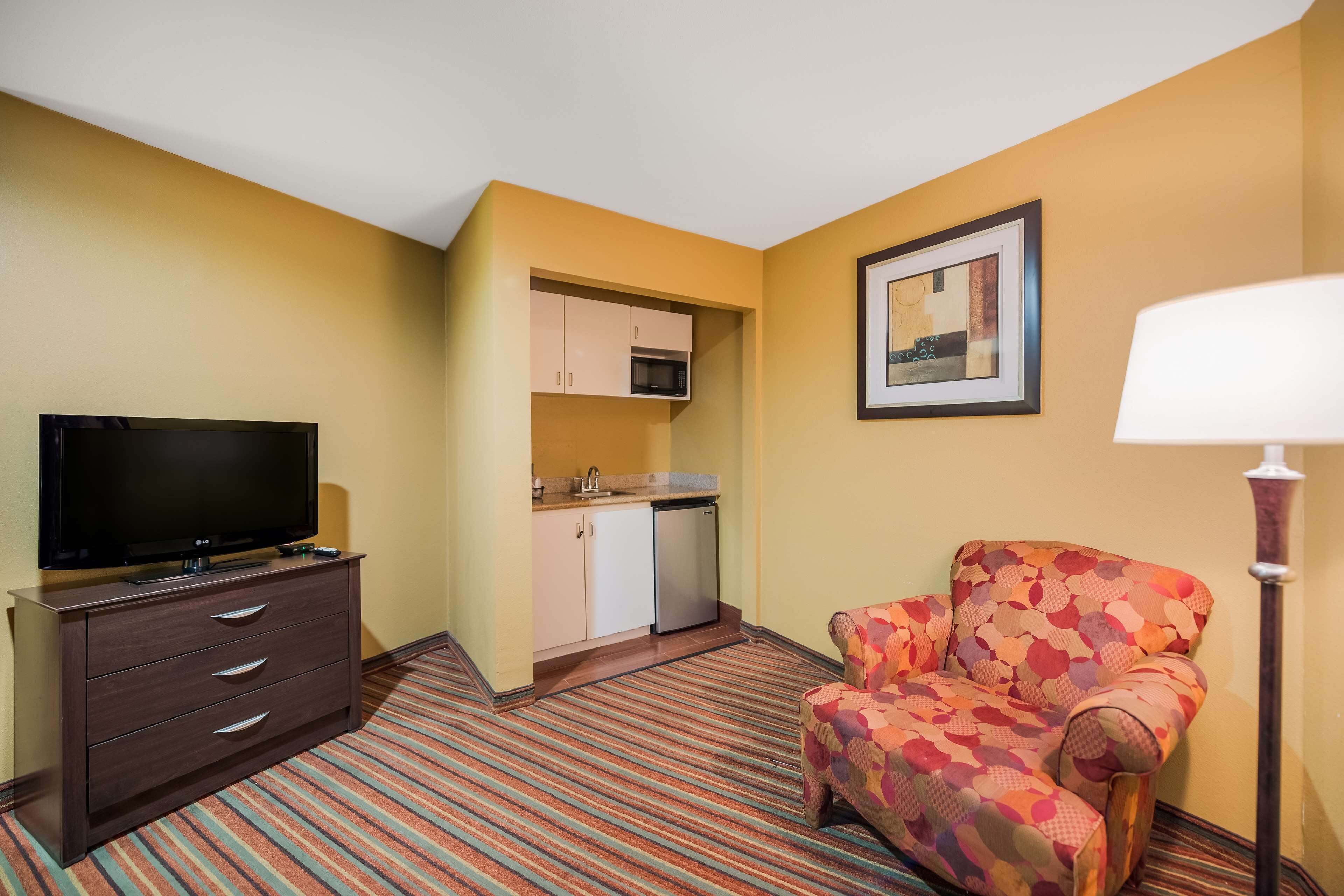 Habitación de hotel con televisión, cómoda, sillón estampado, lámpara, microondas y pequeña cocina.