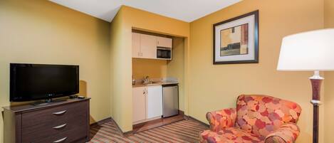 Habitación de hotel con televisión, cómoda, sillón estampado, lámpara, microondas y pequeña cocina.