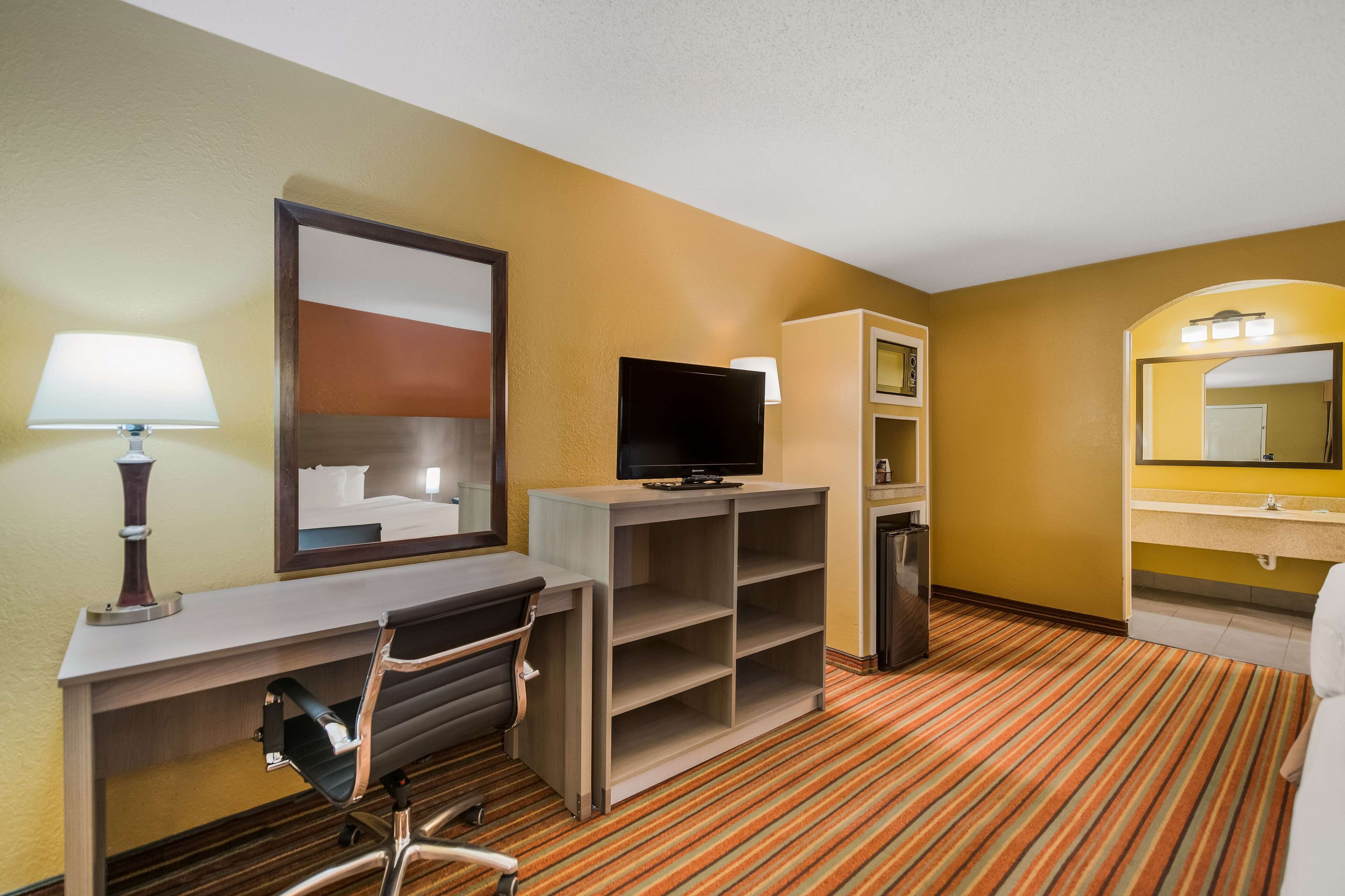 A hotel room with a desk, chair, television, shelves, and a bathroom with a mirror and sink.