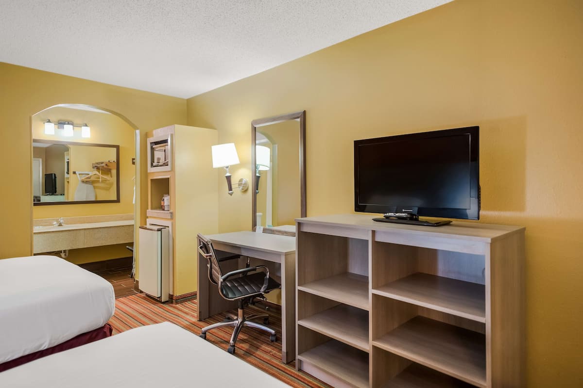 A hotel room with two beds, a desk, a chair, a TV, a mirror, and a shelf.