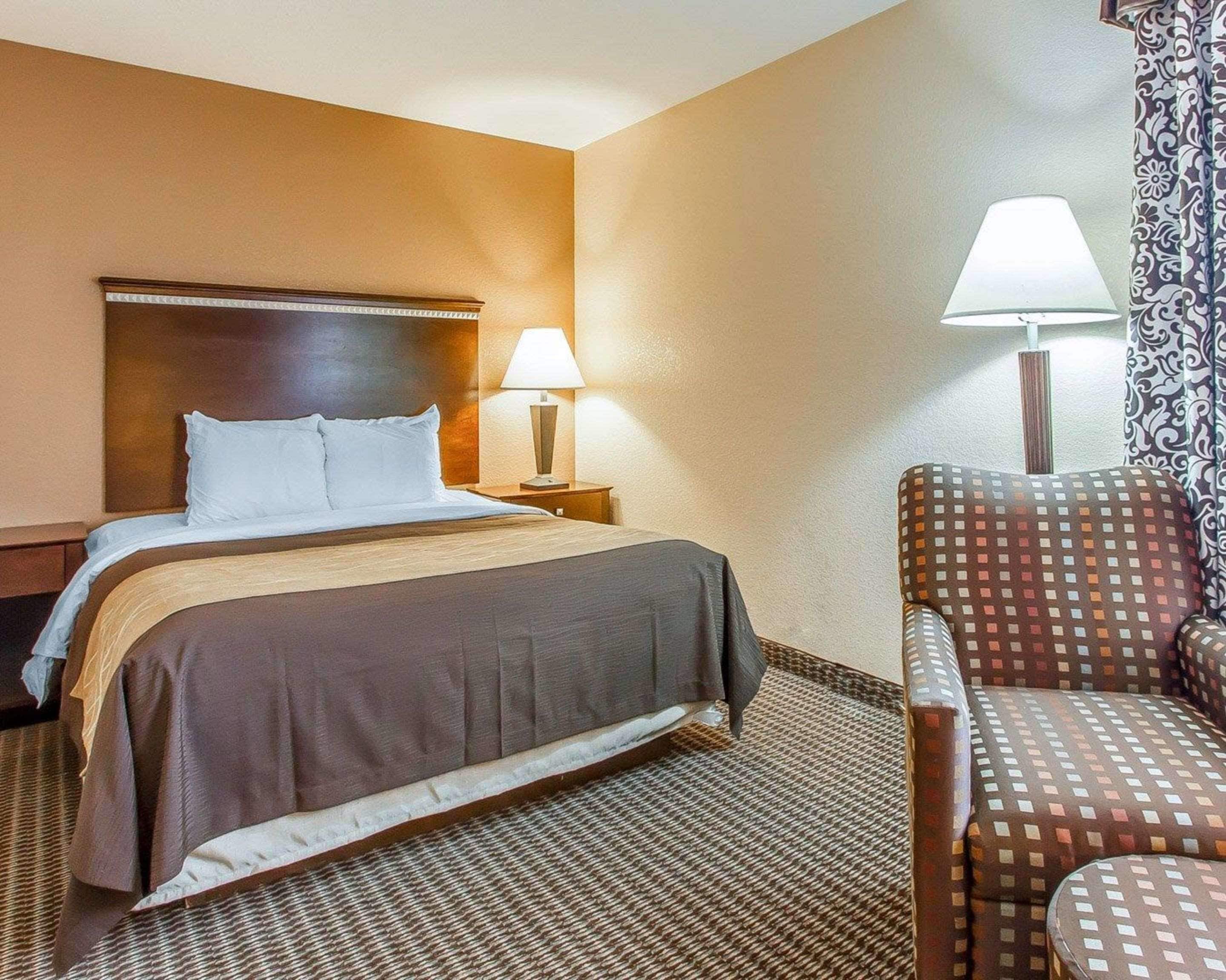 A hotel room with a bed, a nightstand with a lamp, a patterned armchair, and a patterned curtain.