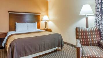 A hotel room with a bed, a nightstand with a lamp, a patterned armchair, and a patterned curtain.