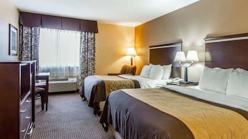 A hotel room with two beds, a desk, and a chair. There is a window with curtains, a lamp, and a wall-mounted phone.