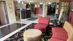 A lobby with a reception desk, red patterned seating, a round beige ottoman, and a potted plant.
