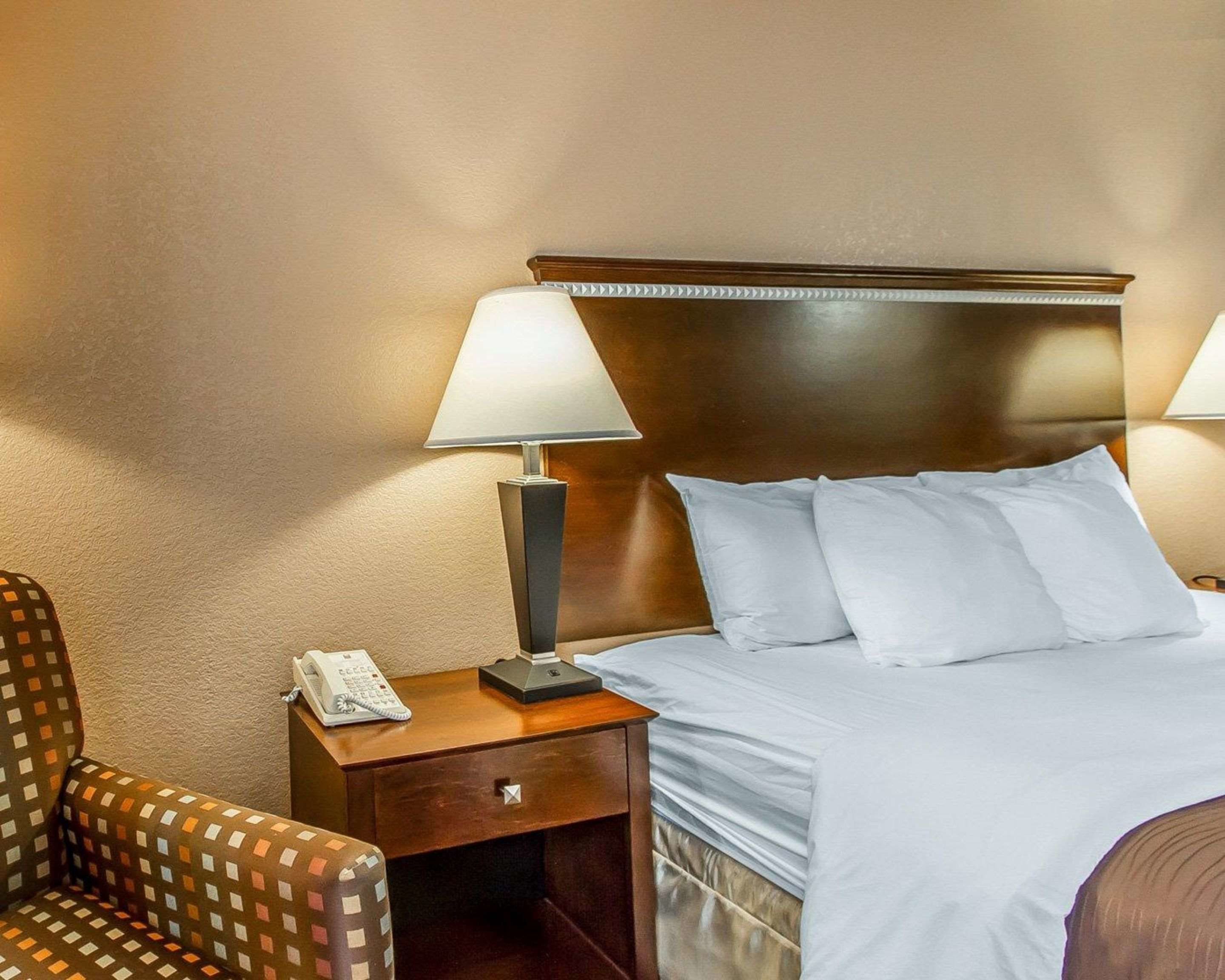 A hotel room with a wooden headboard, a bed with white linens, two bedside lamps, a telephone on a wooden nightstand, and a patterned armchair.