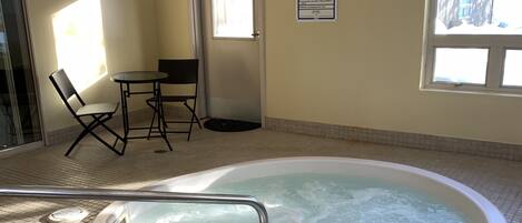Una piscina interior amb un banyera d'hidromassatge, una taula amb cadires i vista cap a l'exterior.