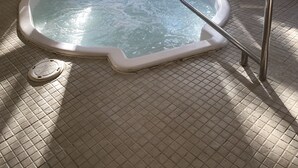 Un jacuzzi en una habitación con suelo de baldosas, dos ventanas con vistas a árboles, y una lámpara fijada a la pared.