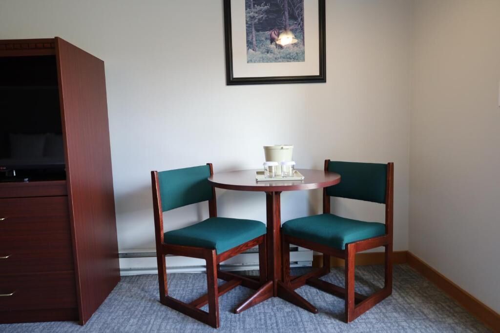 A corner room with a wooden desk, two chairs, a small table with a lamp, and a framed picture on the wall.