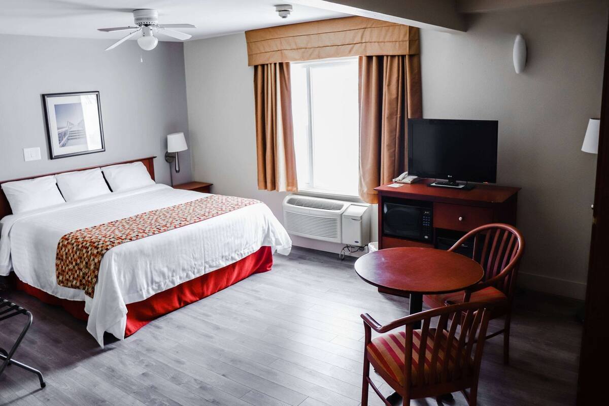 A hotel room with a bed, a desk with a television, a chair, a table, and a window with curtains.