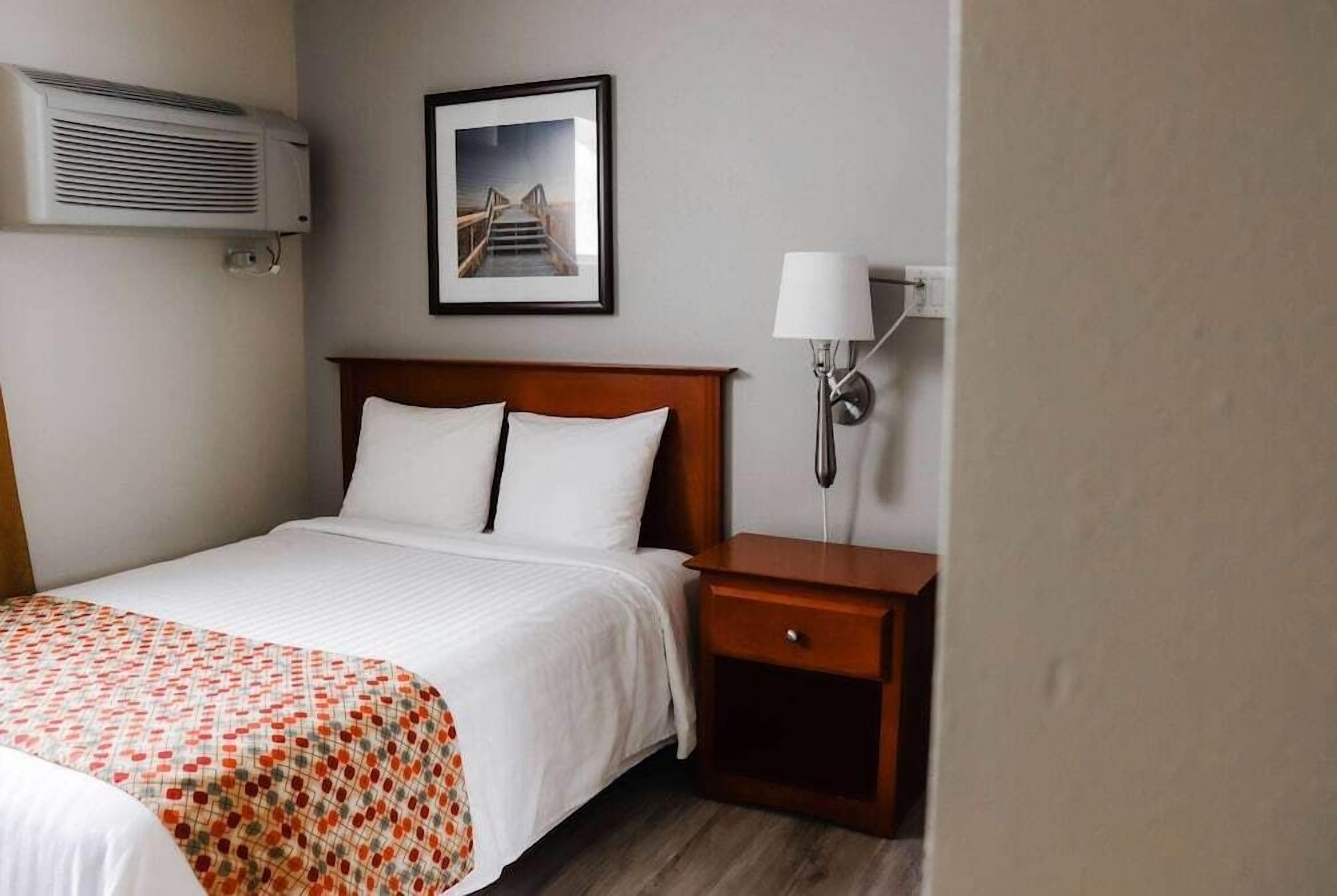 Habitación de hotel con cama, mesita de noche con lámpara, aire acondicionado y un cuadro en la pared.